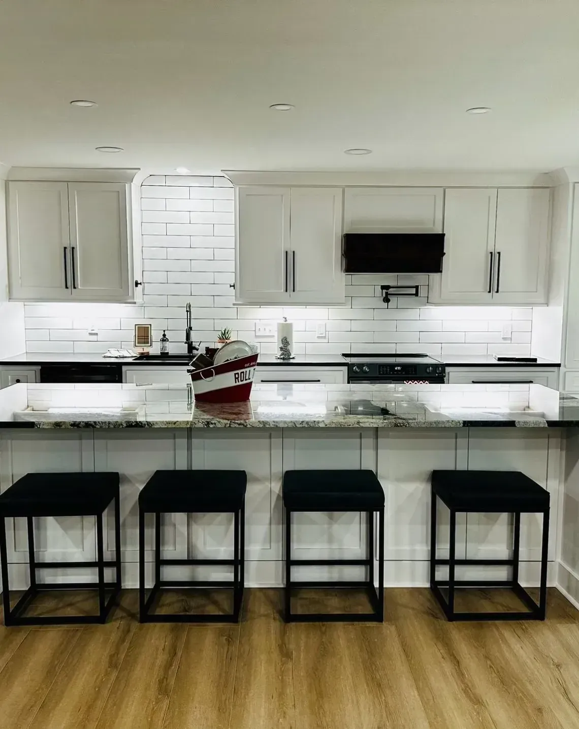 A modern kitchen with white cabinetry, a black refrigerator, a granite-topped island, and four black stools on wood floors.