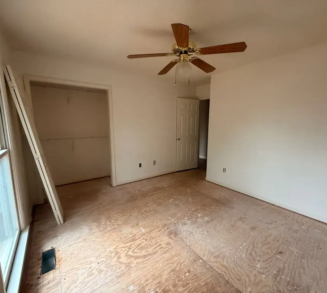 A room under renovation with bare plywood floors, white walls, a ceiling fan, a window, and a leaning closet door.