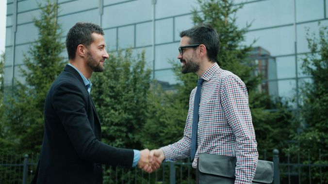 Two men shaking hands outside a glass office building, both in business attire.