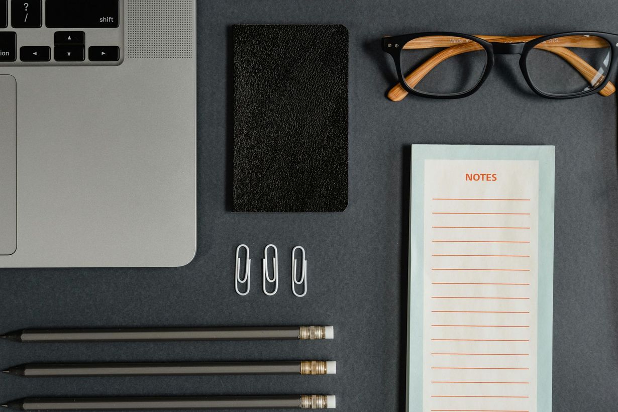 Desk flat lay with laptop corner, glasses, notebook, paper clips, and three pens on a dark surface