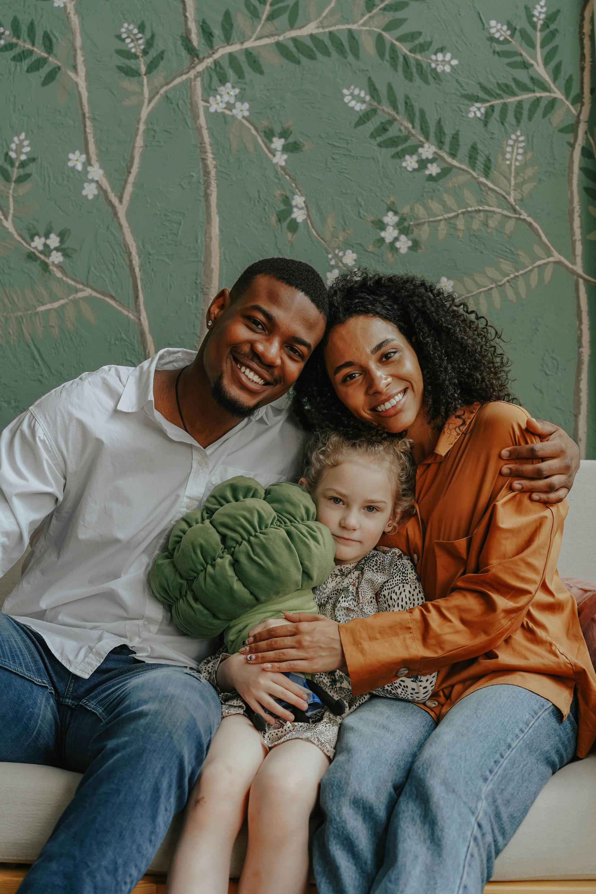 Three people smiling on a couch, hugging a green cushion, with leafy wallpaper behind them.