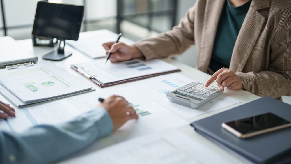 People reviewing documents at a desk with a laptop, calculator, and tablet in a bright office