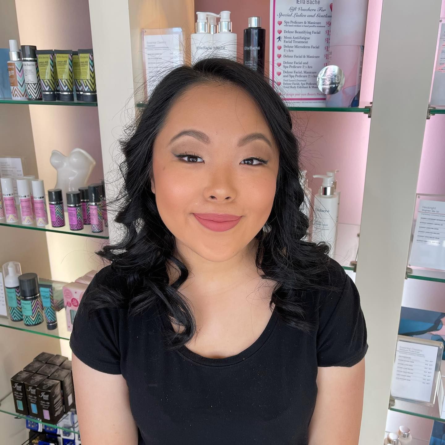 Woman with black hair smiles, standing in front of beauty products displayed on shelves.