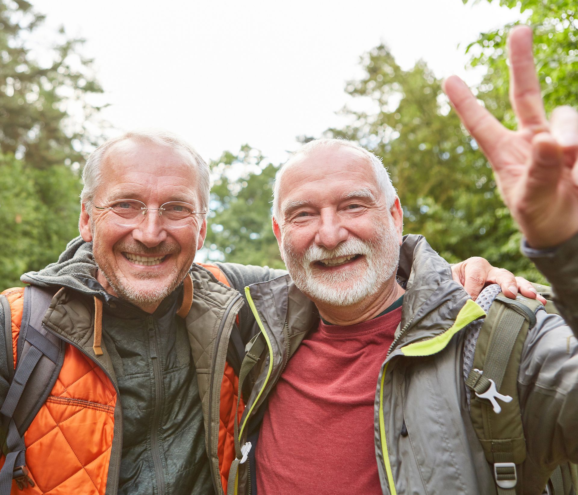Two older men smiling, one giving a peace sign, outdoors in a forest.