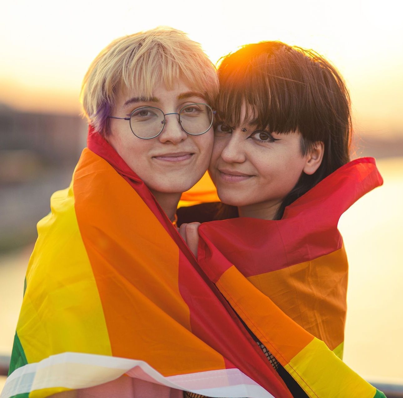 Two people wrapped in a rainbow flag, smiling in front of a sunset.