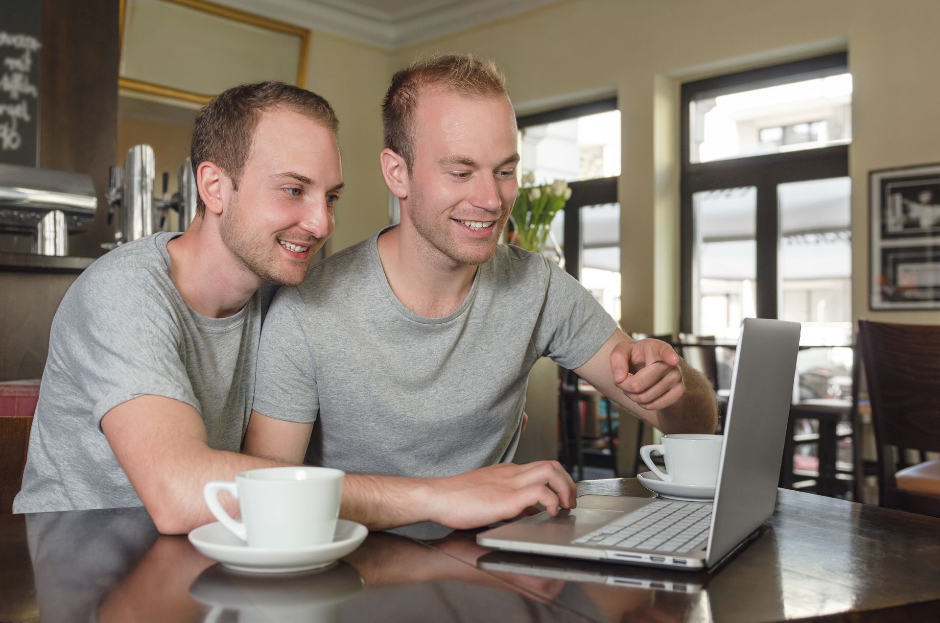 Two men looking at a laptop in a cafe, smiling. Cups of coffee on the table.
