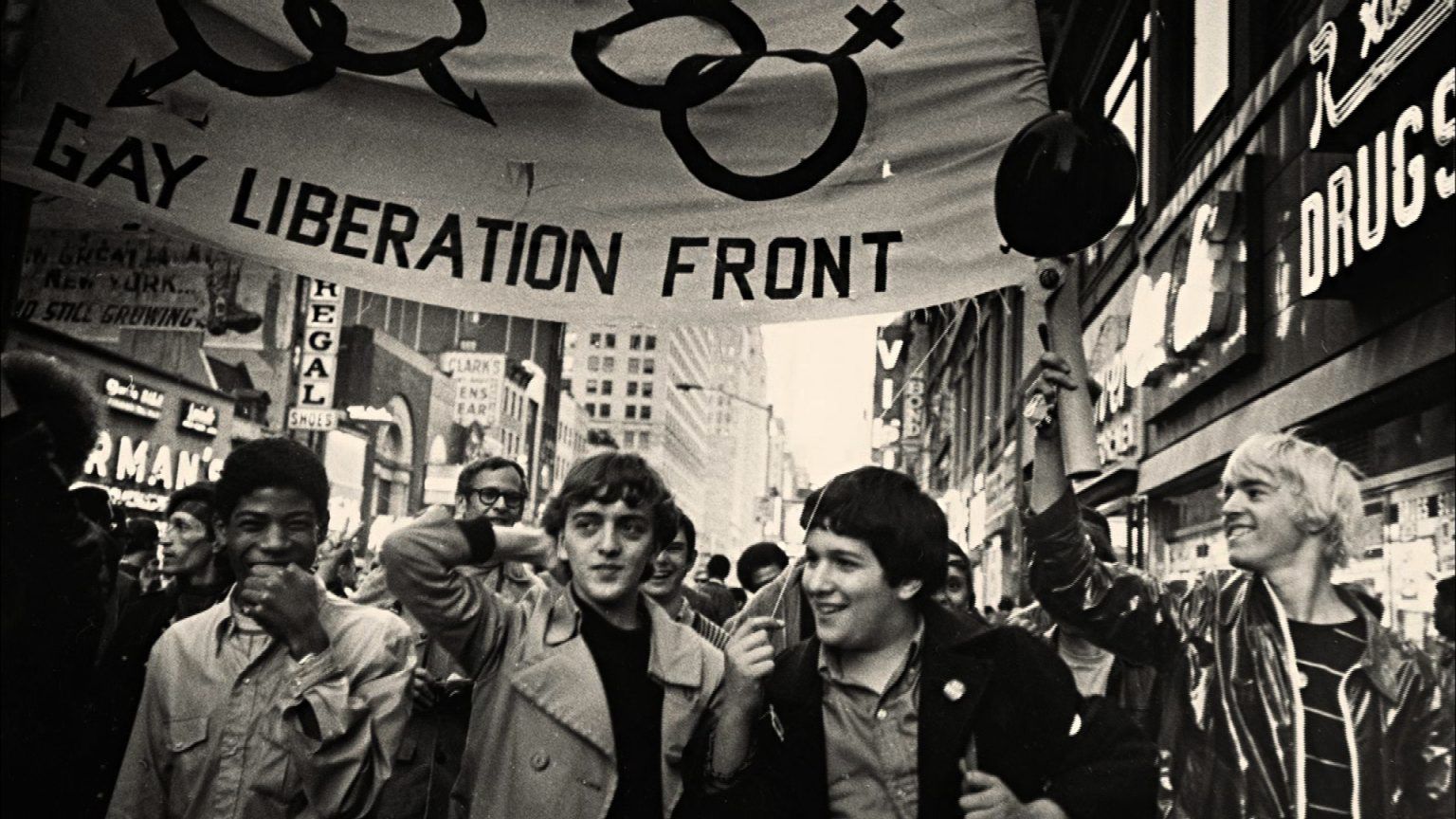 Black and white photo of a Gay Liberation Front march. People carry a banner, walking down a city street.