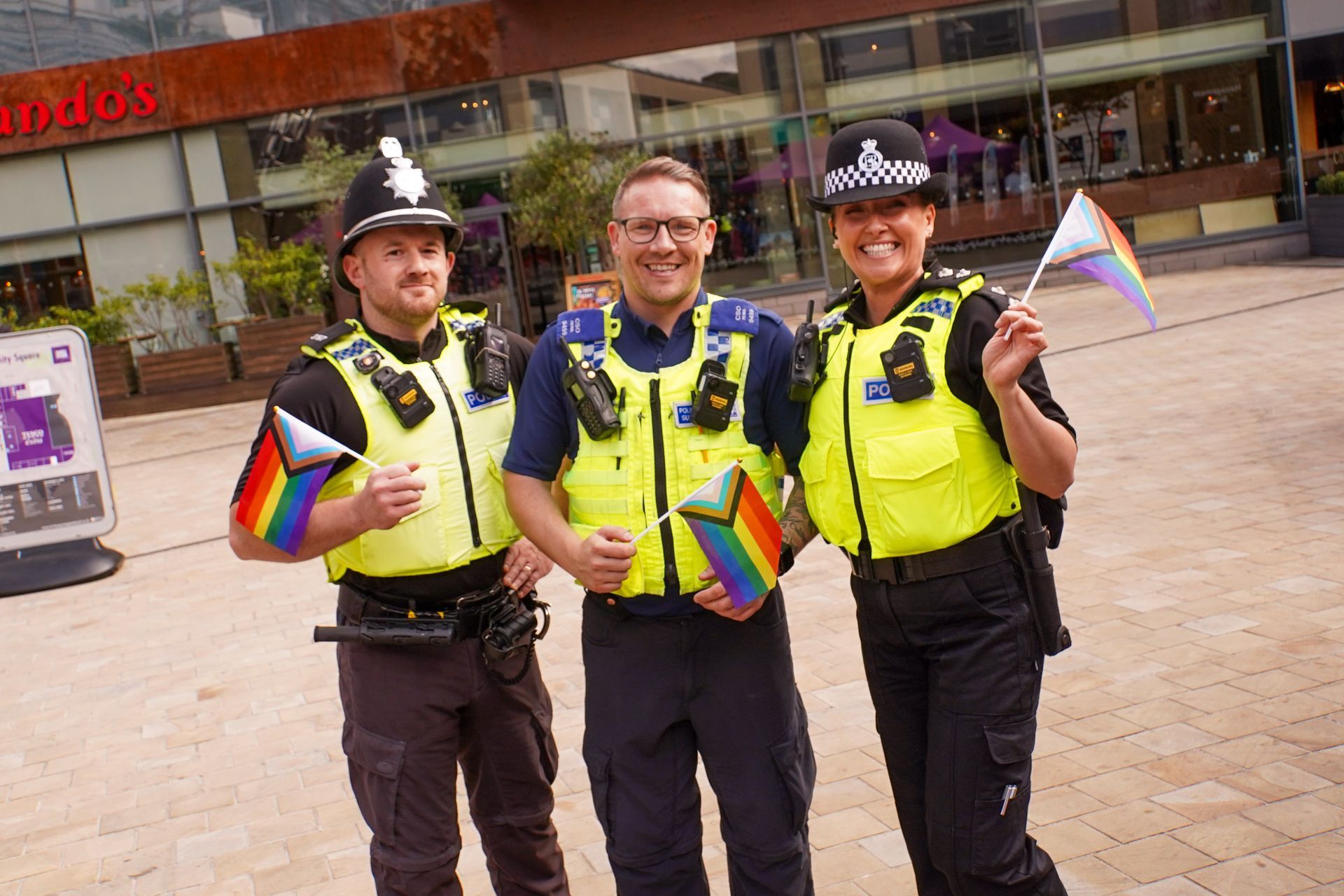Three police officers smiling, holding rainbow flags. Building exterior in background.