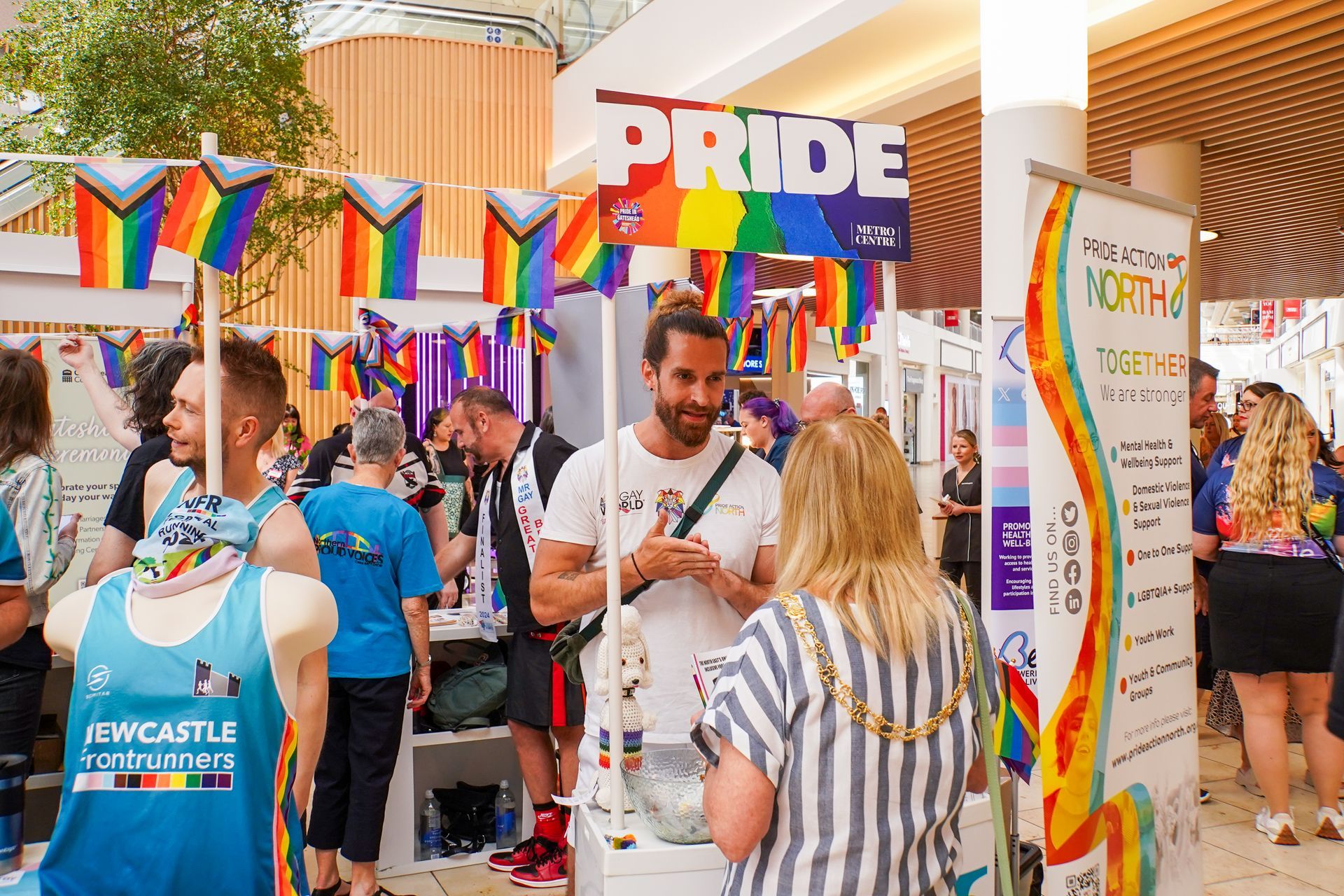 Pride event booth with rainbow flags and merchandise; people interact, shopping and talking.