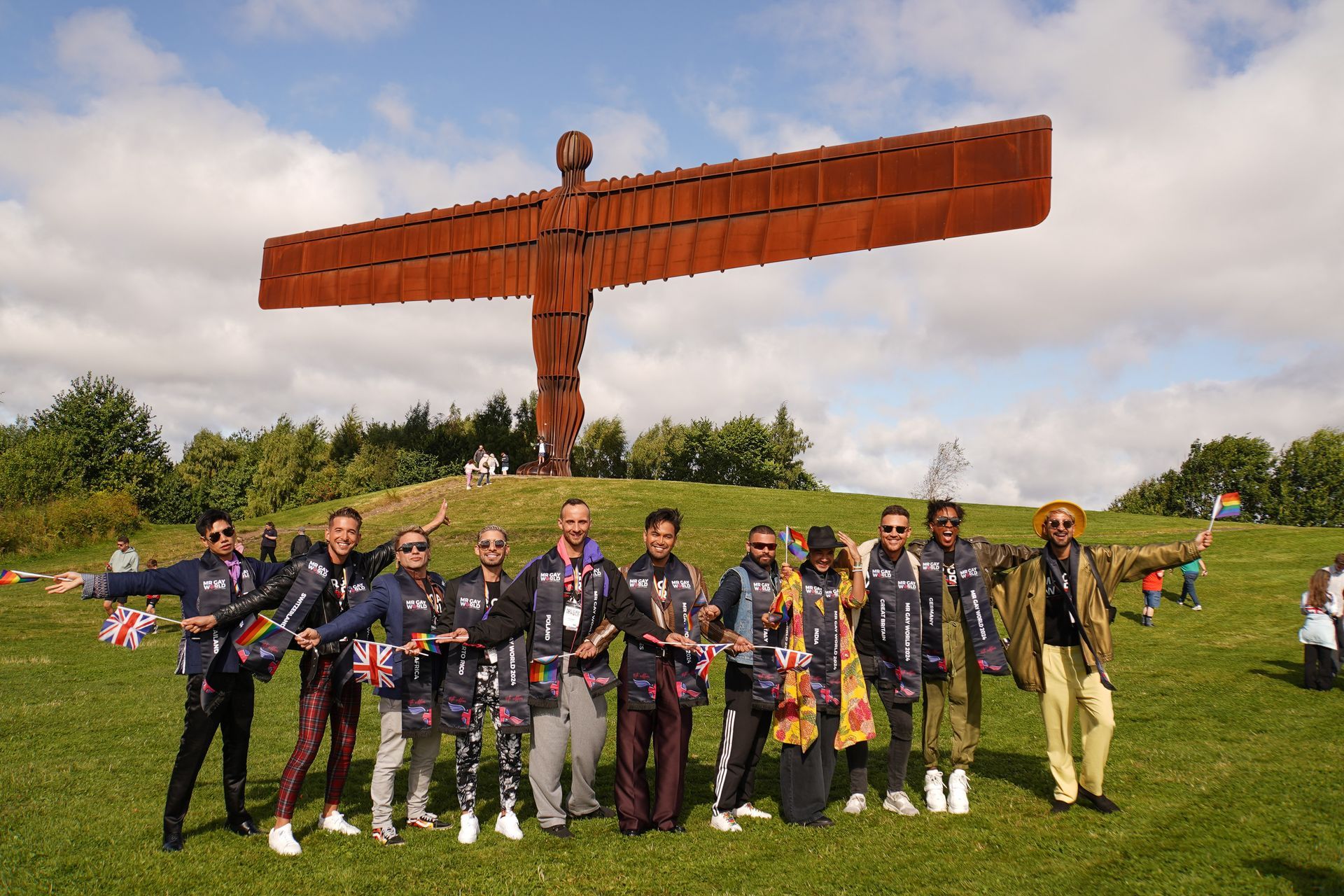 Group of people standing in front of the Angel of the North sculpture, arms outstretched. Green field, blue sky.