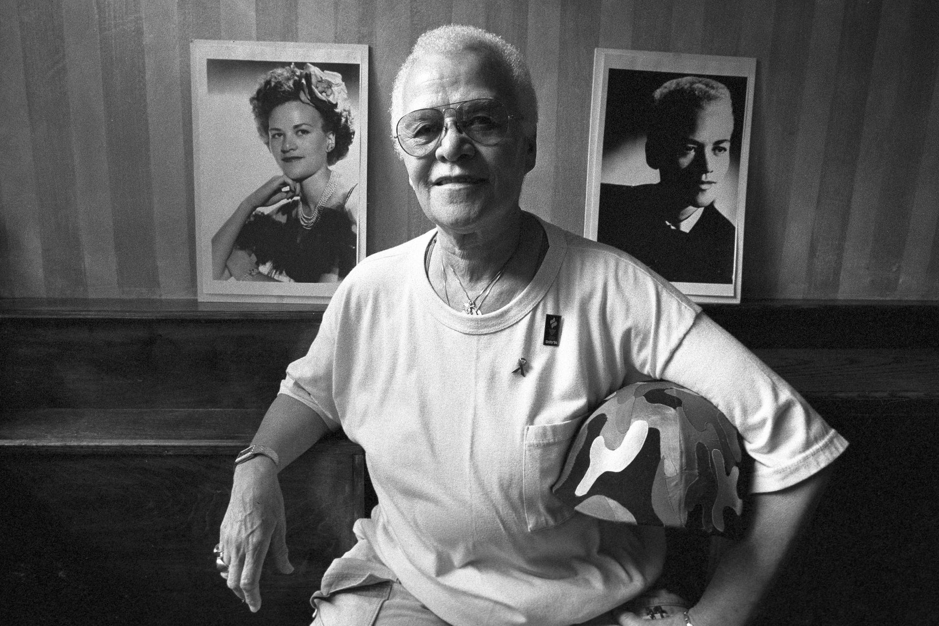 Woman seated, holding a camouflage helmet, with portraits on wall behind.