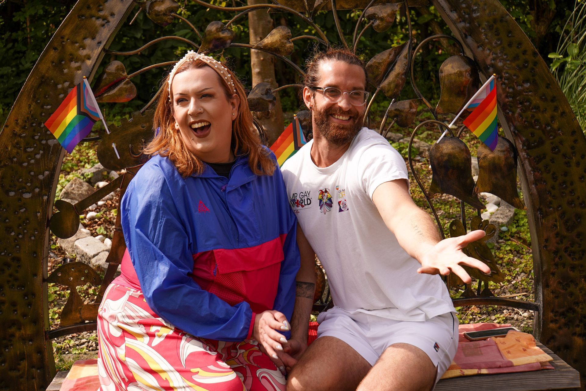 Two people smiling with rainbow flags; sitting outdoors.