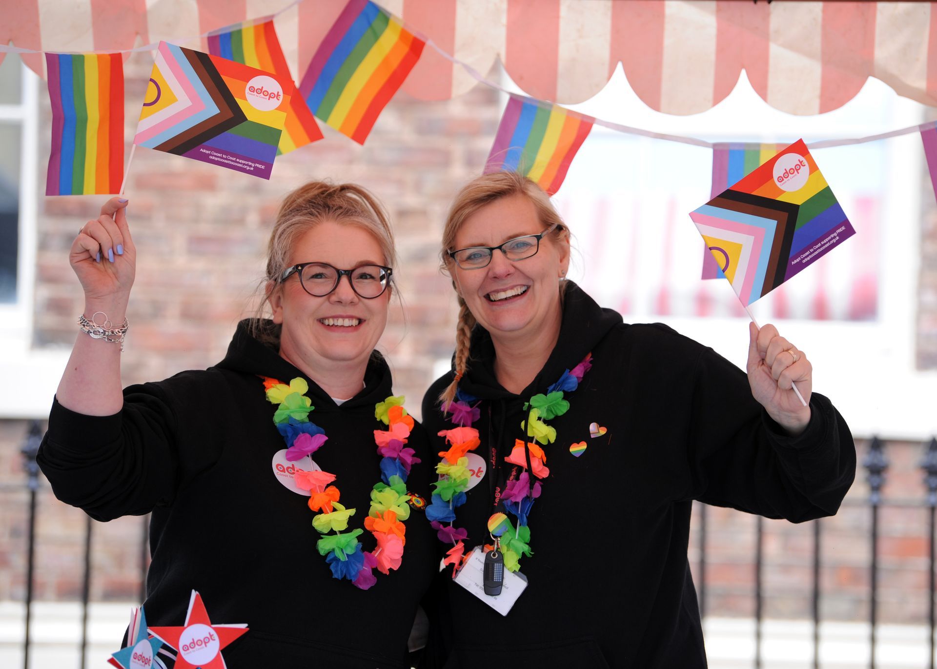 Two women smiling, waving Pride flags, under a rainbow banner.