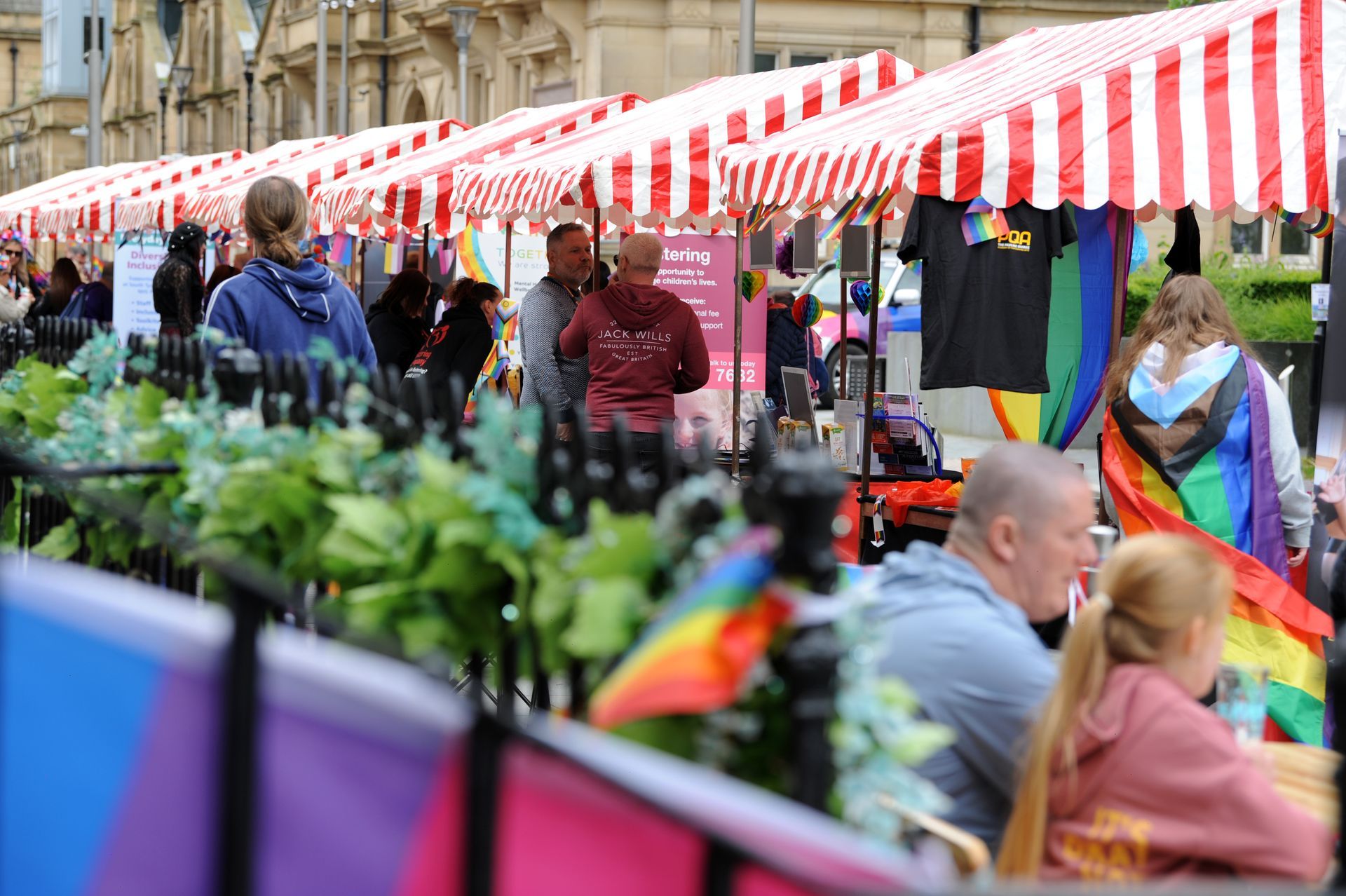 Pride market with rainbow flags, striped canopies, and people browsing stalls.