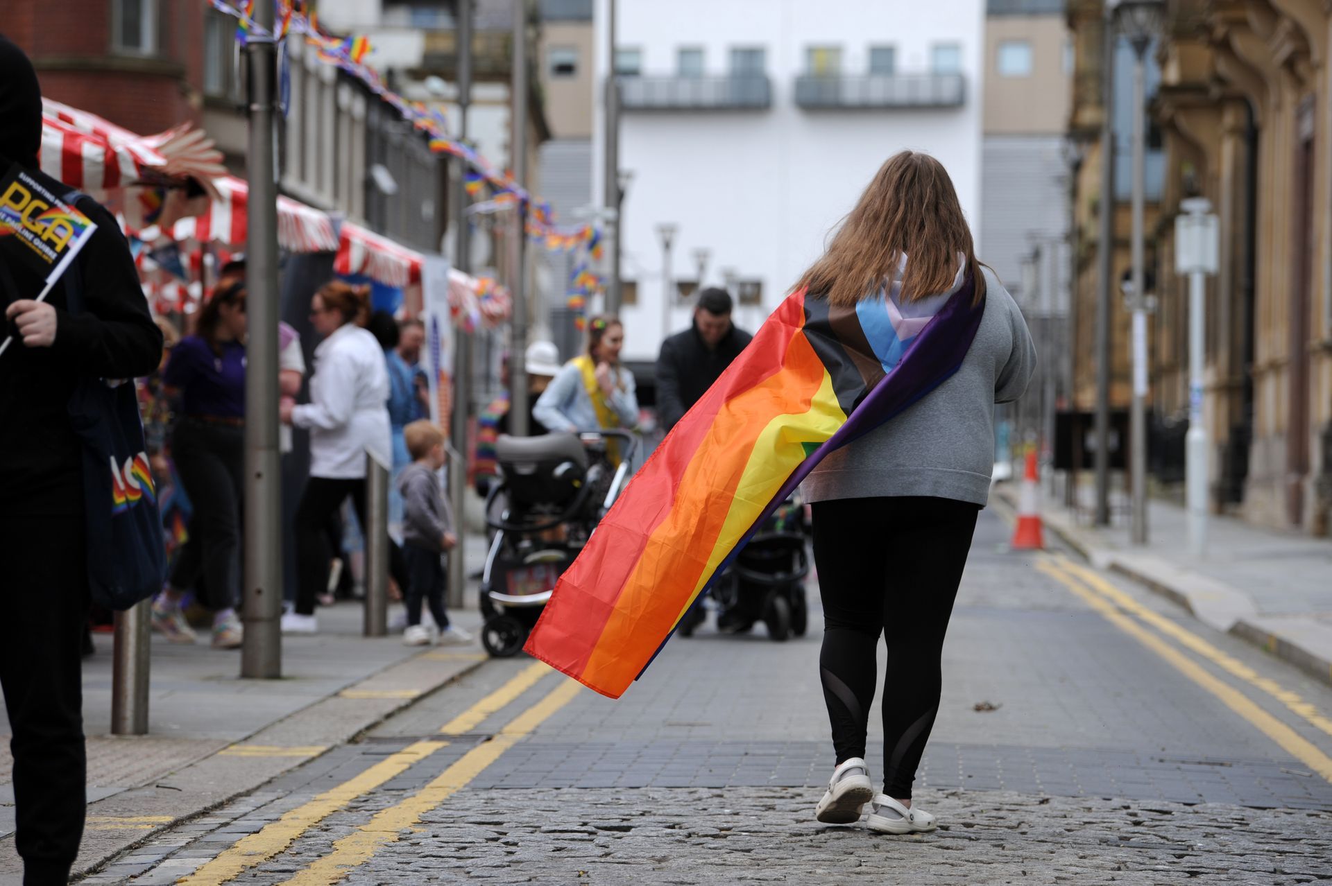 Person walking down a street, holding a rainbow flag. People and buildings in the background.