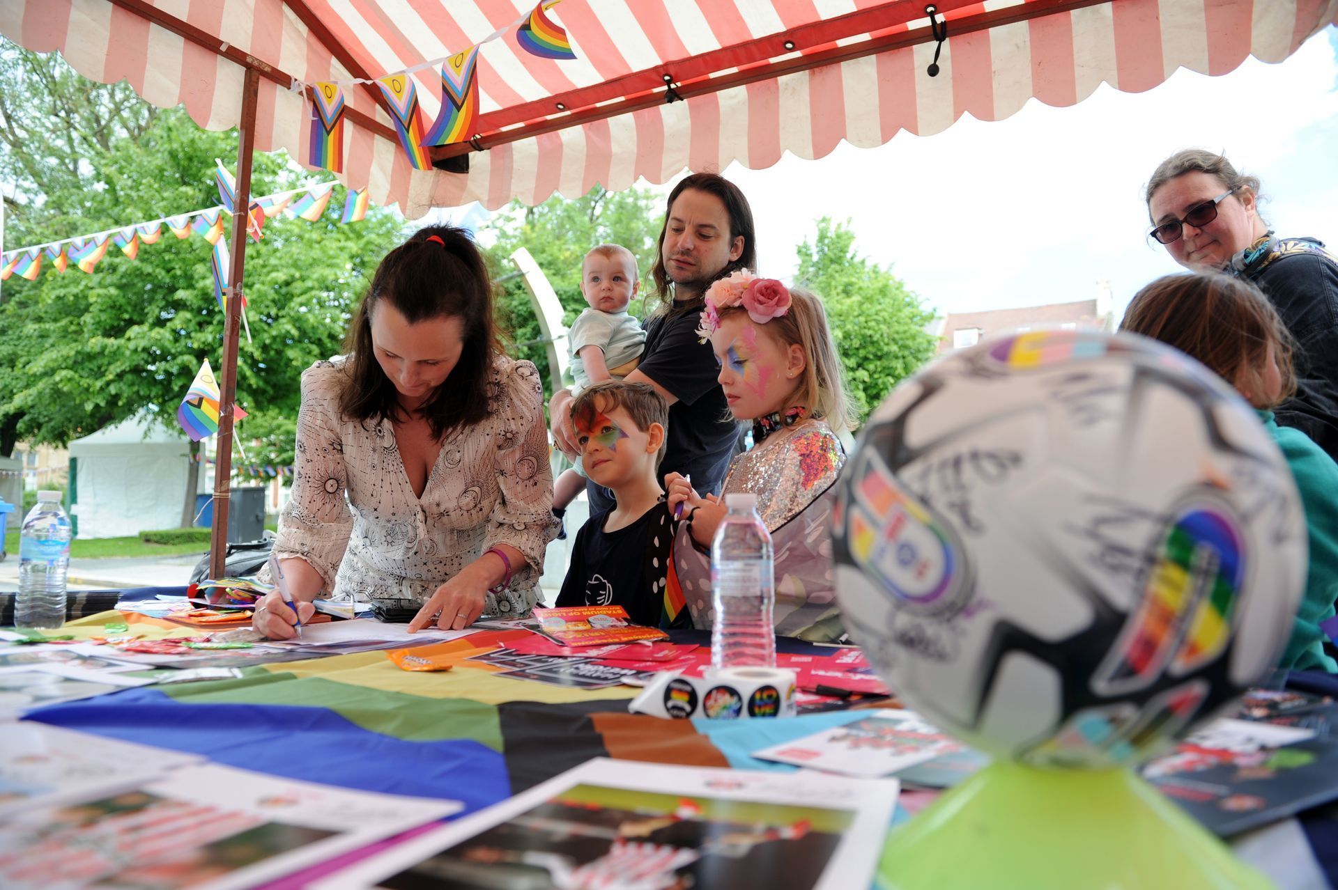 People at a table under a striped tent, drawing and decorating a soccer ball with rainbow colors.