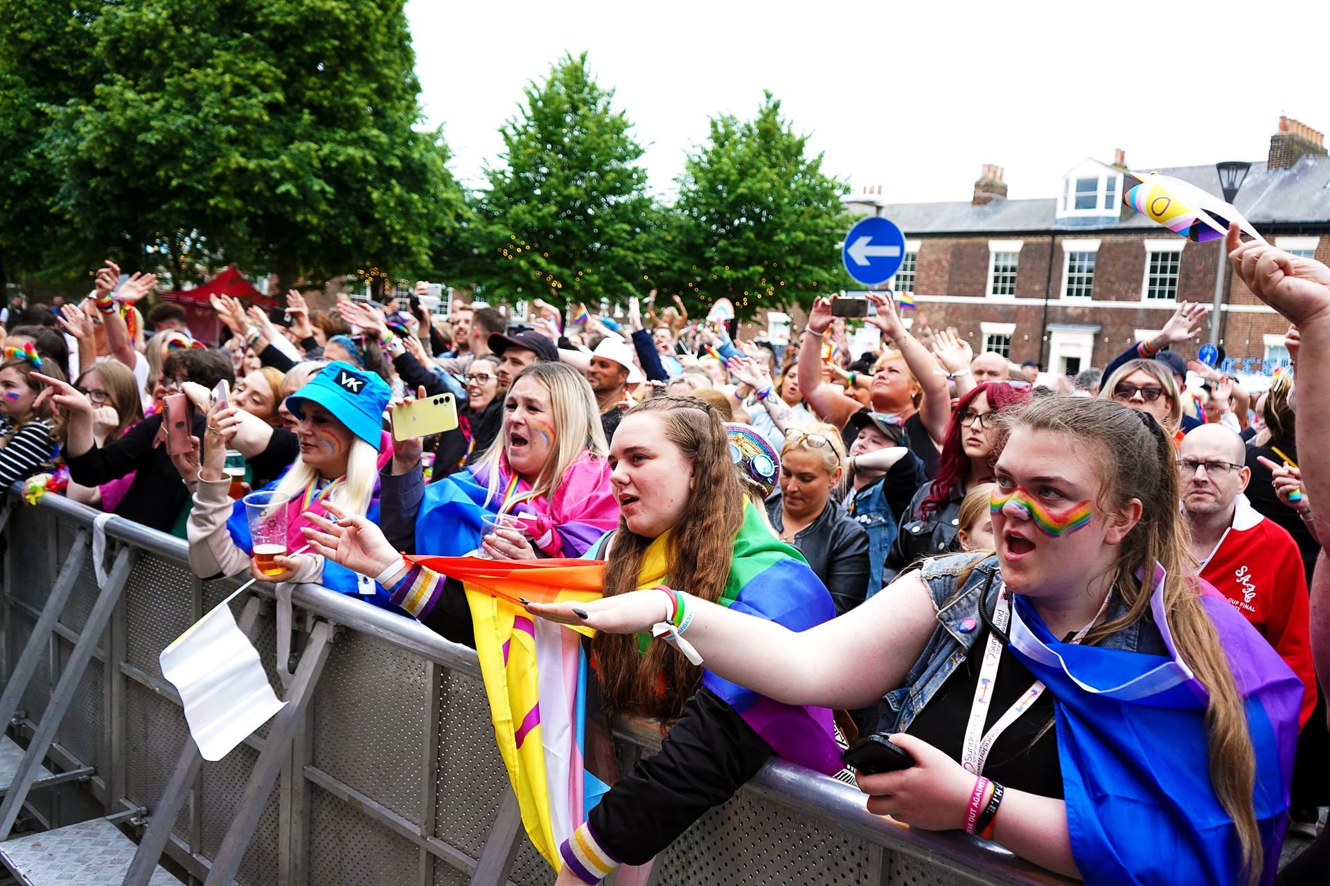 People cheering at an outdoor Pride event, waving flags and holding phones. Rainbow colors are prominent.
