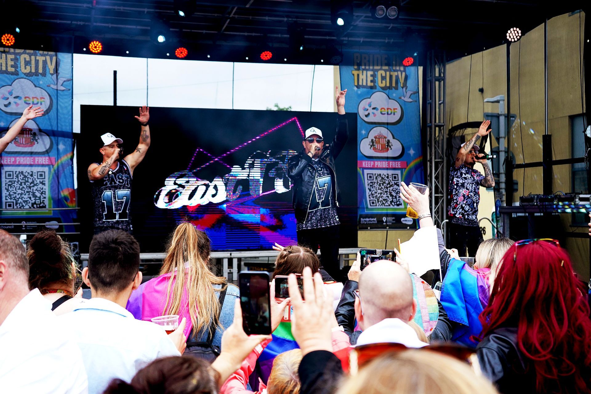 Band performs on stage at an outdoor event. People in the audience record with phones, raise hands.