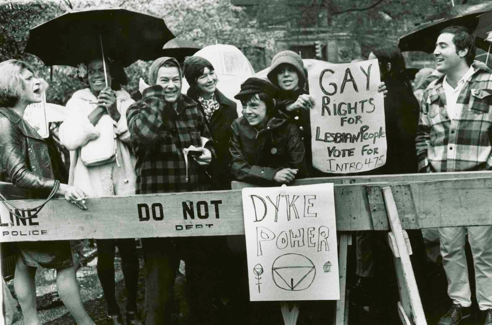 People holding signs advocating for gay rights at a protest, standing behind a barricade.