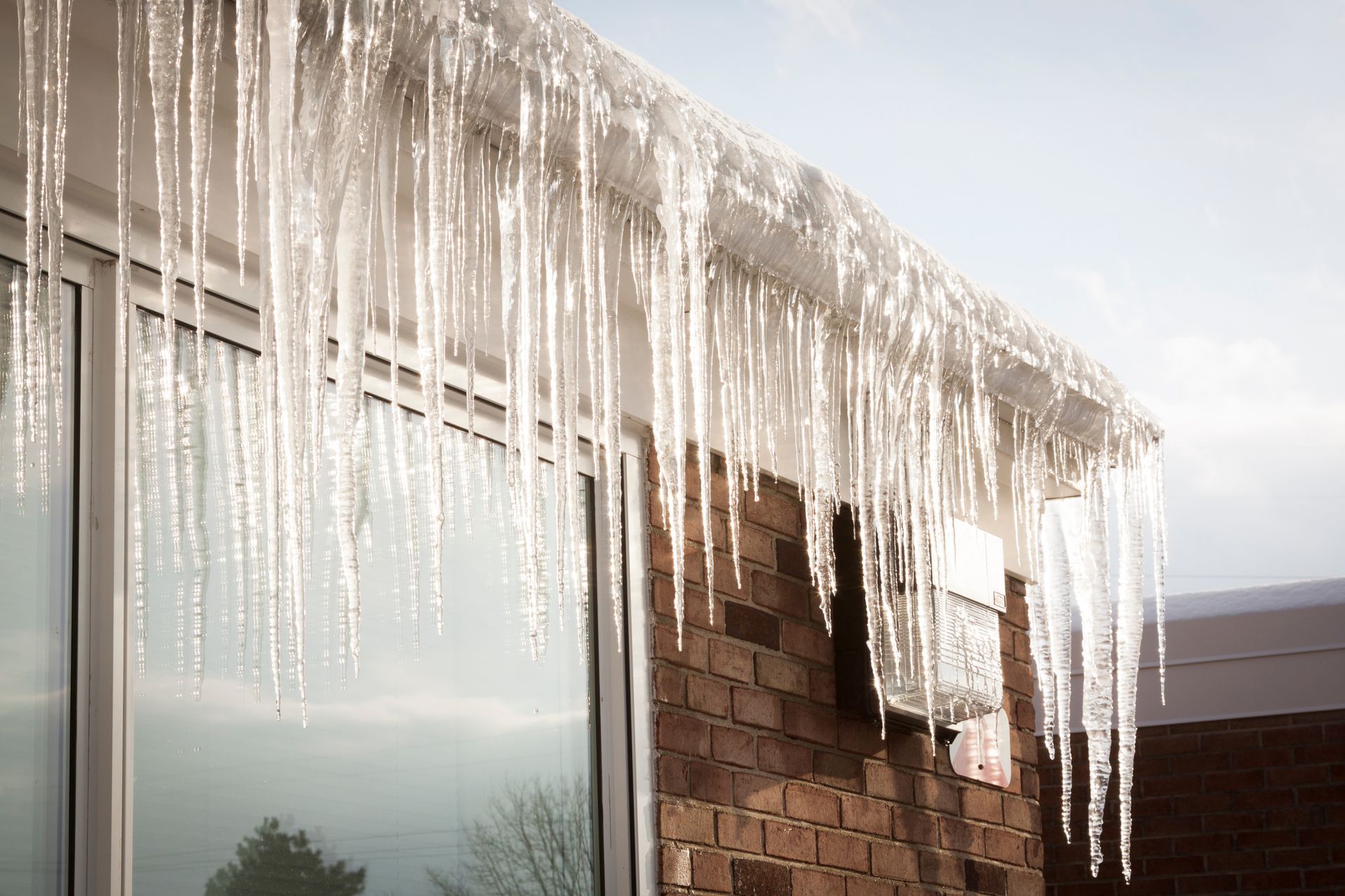 Long icicles hanging from the edge of a house roof above a window and brick wall