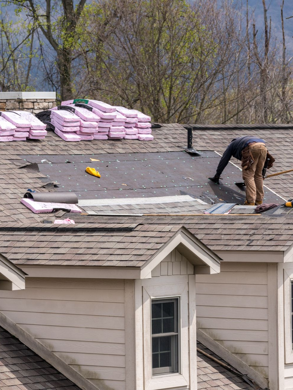 Worker installing shingles on a residential roof with stacks of roofing materials nearby