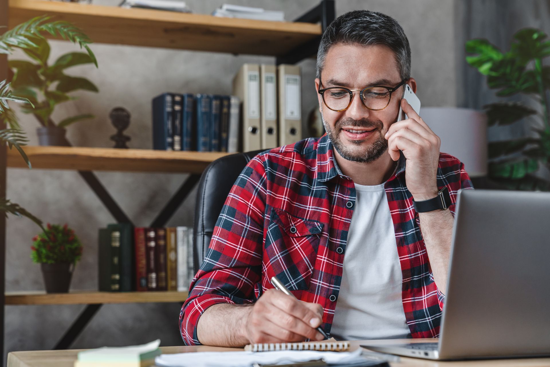 Man in a red plaid shirt on a phone call, writing in a notebook beside a laptop at a home desk.
