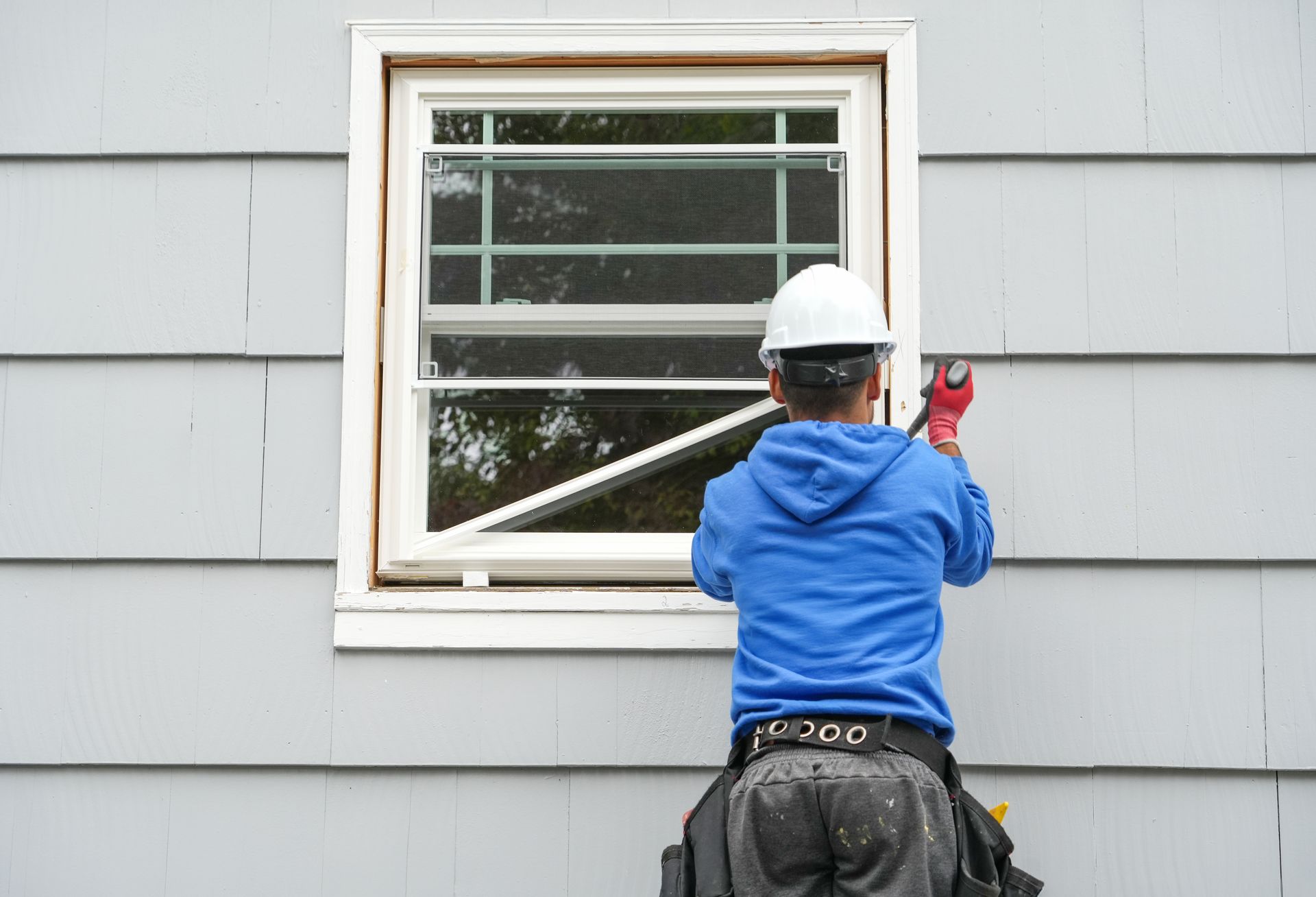 Worker in blue hoodie caulking a white-framed window on a gray house.