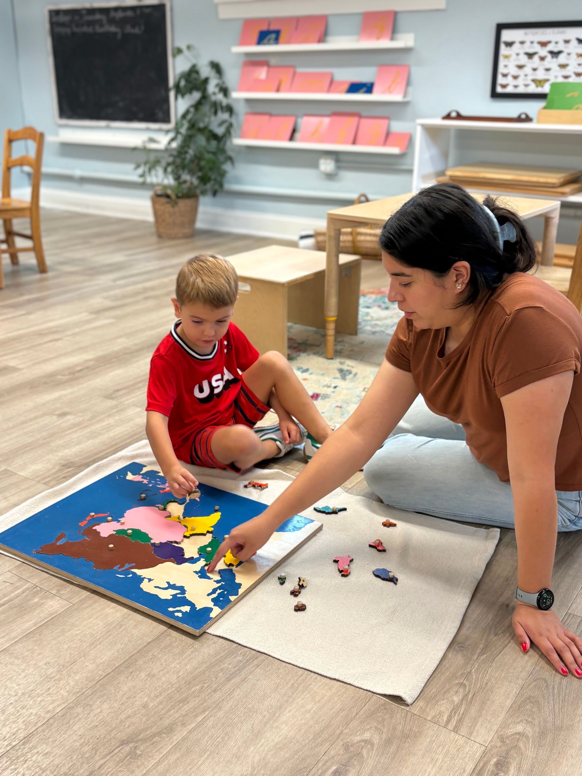 Adult and child kneel by a puzzle map, placing pieces in a bright classroom.