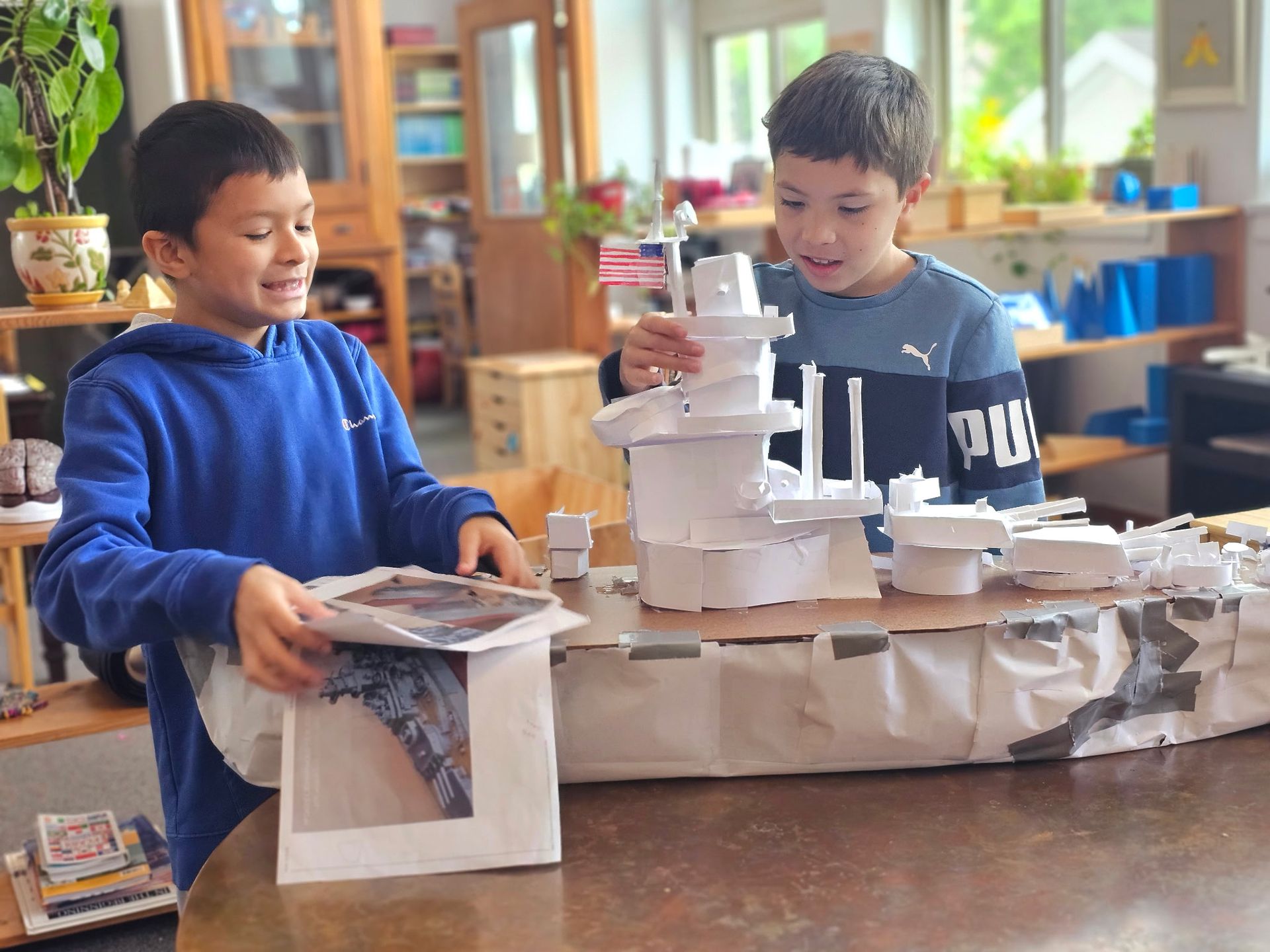 Two children building a cardboard model together in a classroom, one holding a magazine, one smiling.