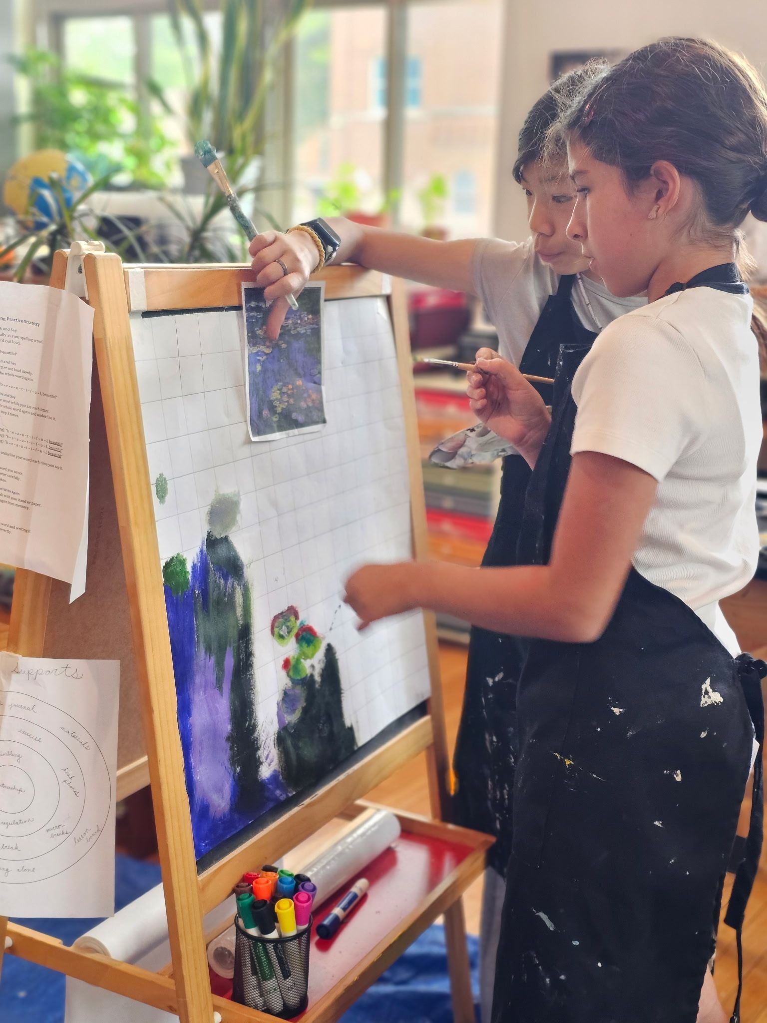 Two children paint at an easel in a bright classroom, adding blue and purple colors to a canvas.