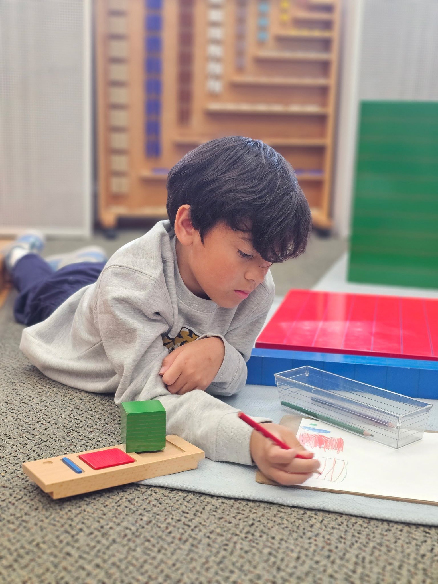 Child lying on the floor, drawing with a red pencil beside colorful blocks and a notebook.