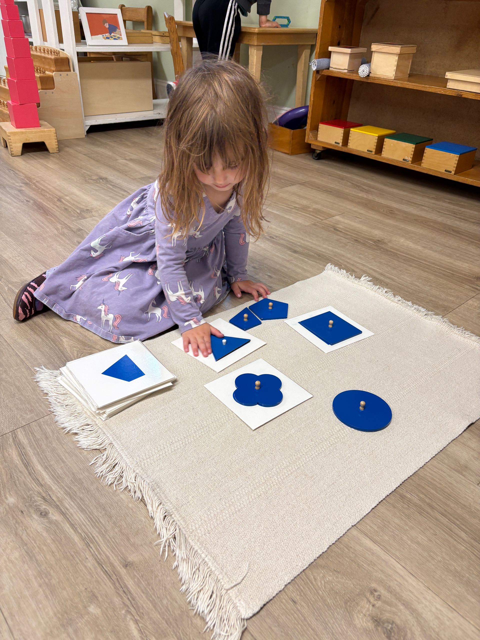 Child kneeling on a rug, matching blue shapes to white cards in a classroom.