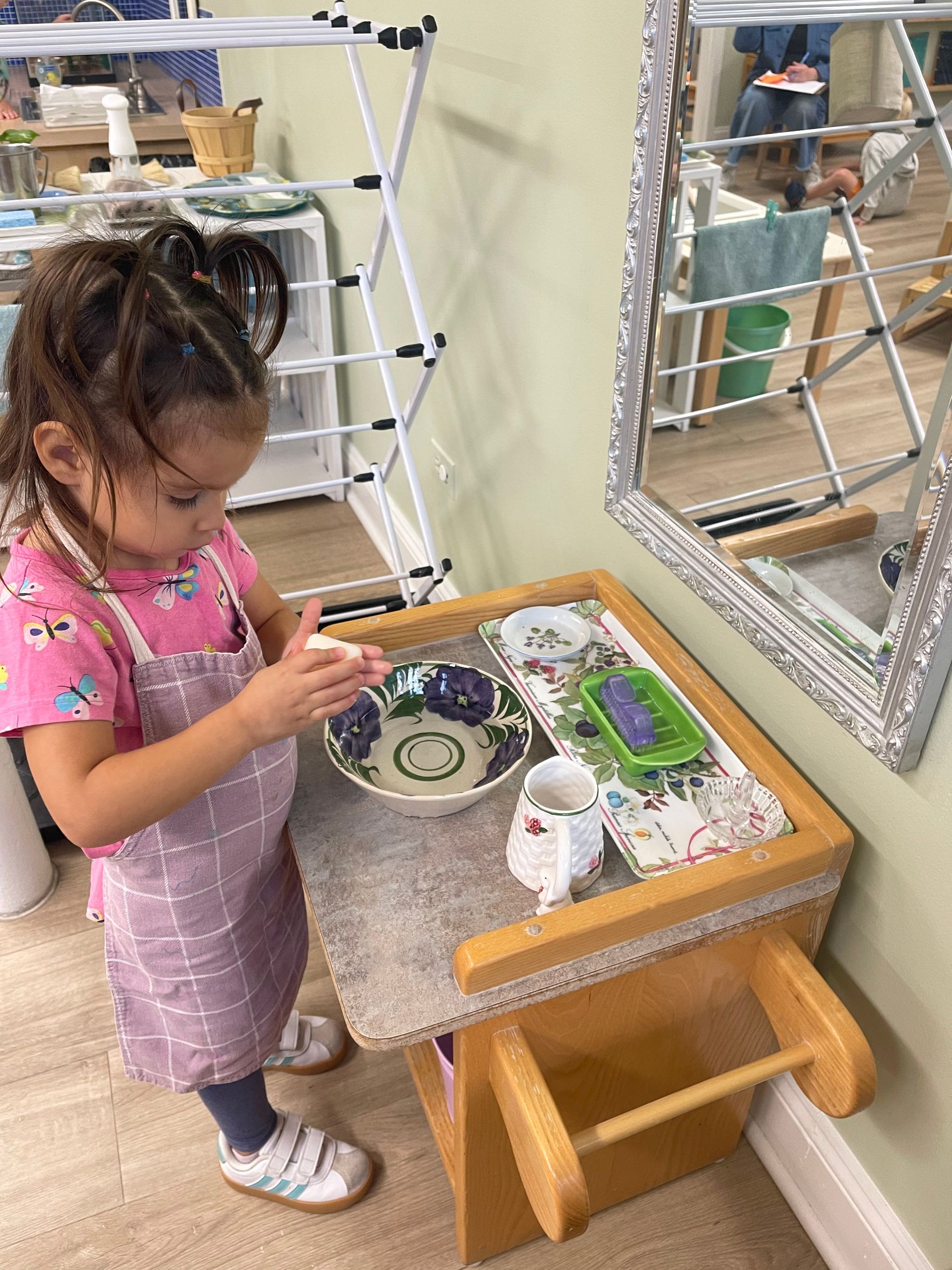 Child in a pink shirt washes hands at a small wooden table beside a mirror.