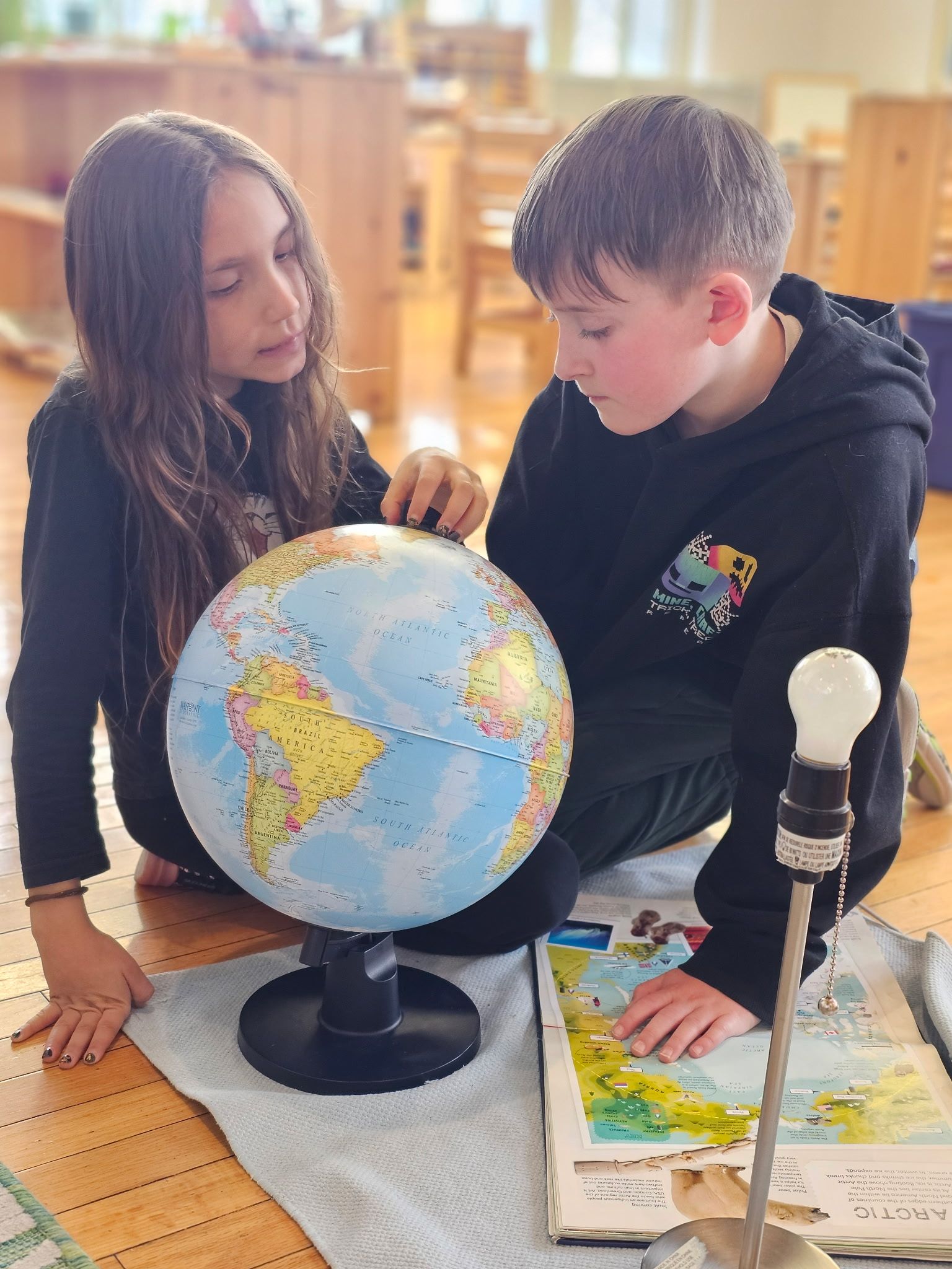 Two children studying a globe together on the floor with books and a magnifying glass