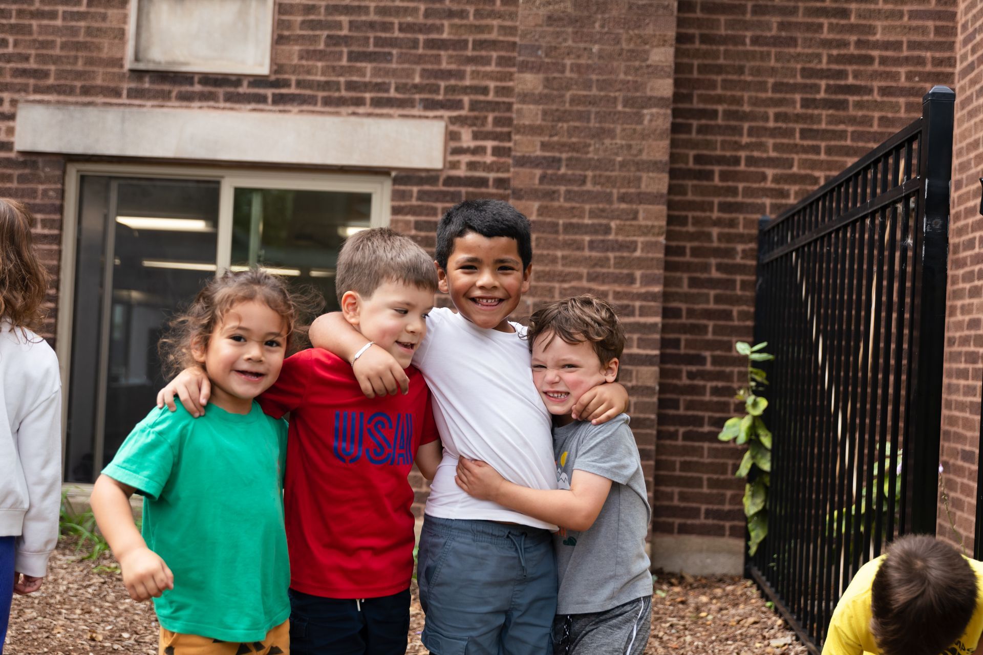 Four children smiling and hugging outside a brick building near a black fence