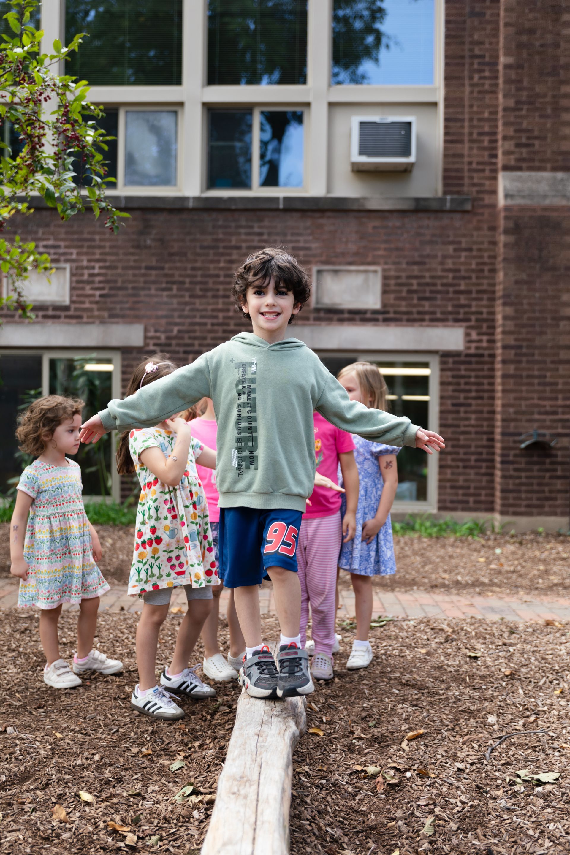 Children balancing on a log outdoors in a playground, with a brick building in the background.