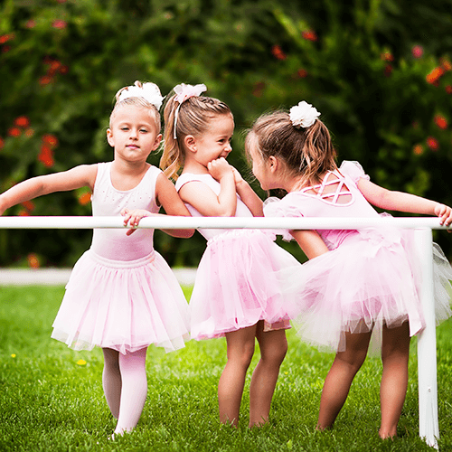 Three children in pink ballet outfits holding a white barre outdoors on grass