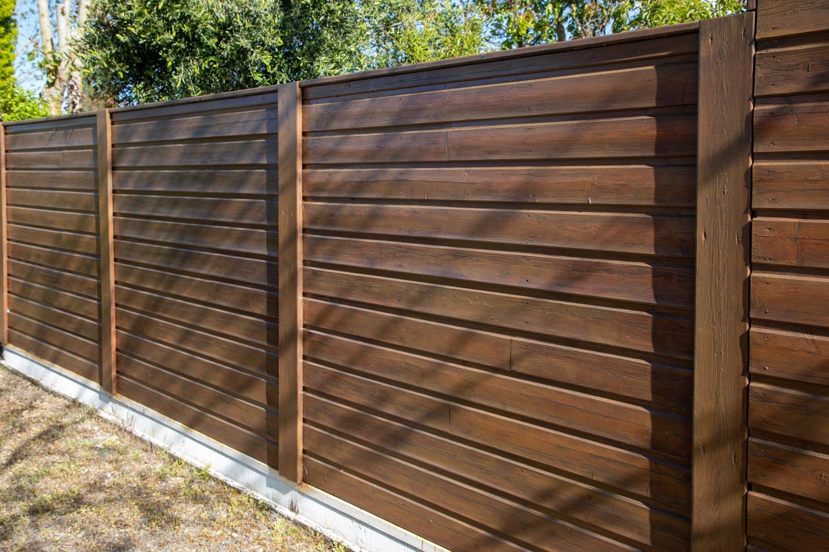 Brown horizontal wooden fence in a yard, with a concrete base, and greenery in background.