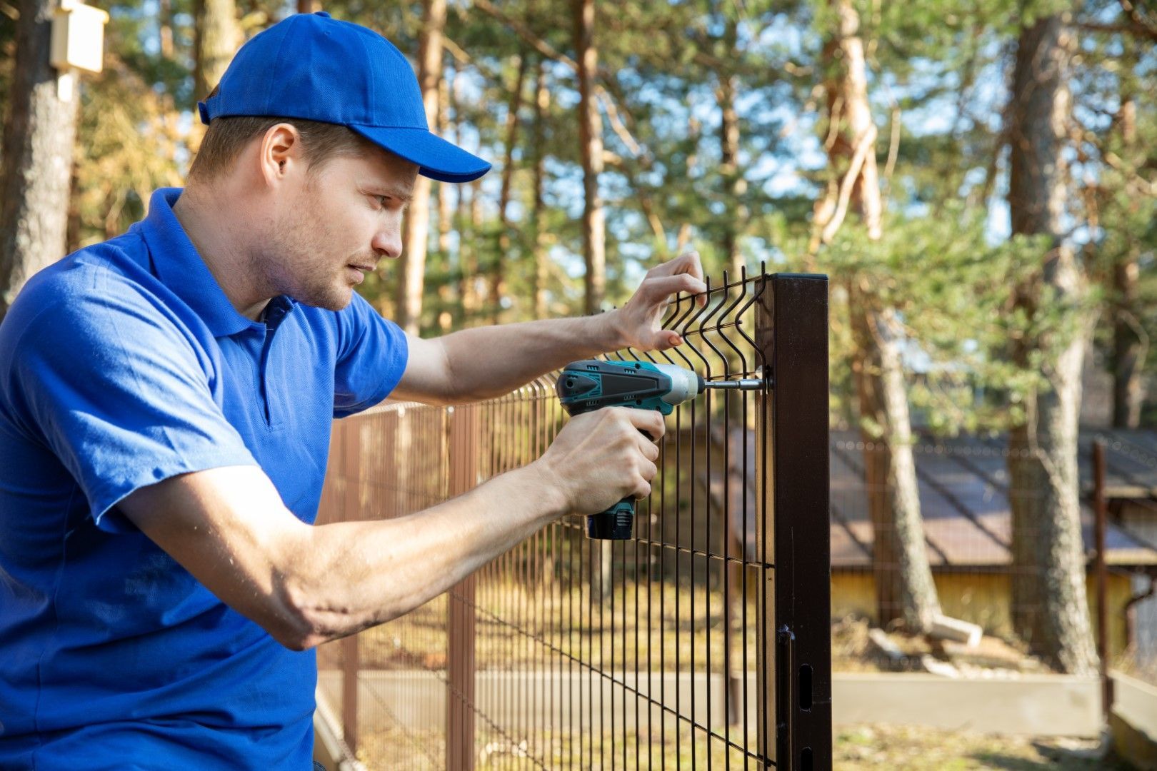 Man in blue uniform using a drill to install a brown metal fence. Outdoors, sunny.