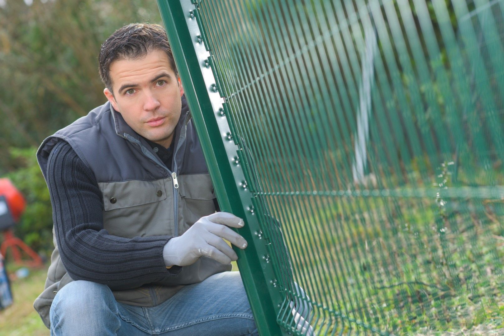 Man in gloves installing a green metal fence outdoors, looking at the camera.