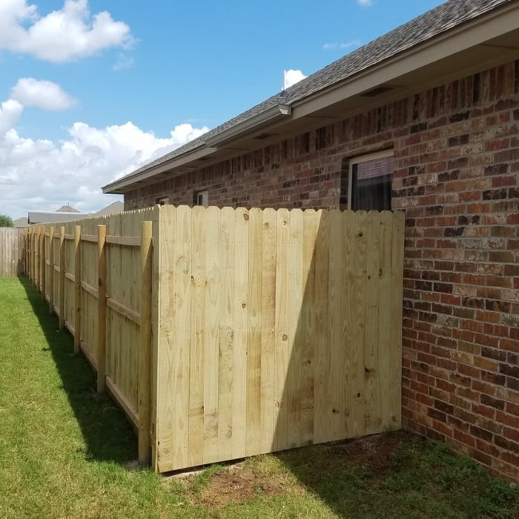 Wooden privacy fence along a brick house beside a grassy yard under a partly cloudy sky