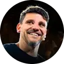 Homem sorridente de camisa escura contra um fundo escuro, iluminado por luz quente de palco.