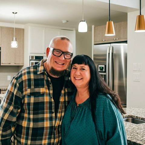 Couple smiling in a modern kitchen. Man wearing glasses and plaid shirt, woman in teal top. Pendant lights overhead.