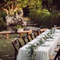 Long outdoor dining table with white tablecloth, greenery centerpiece, and black chairs in a garden setting