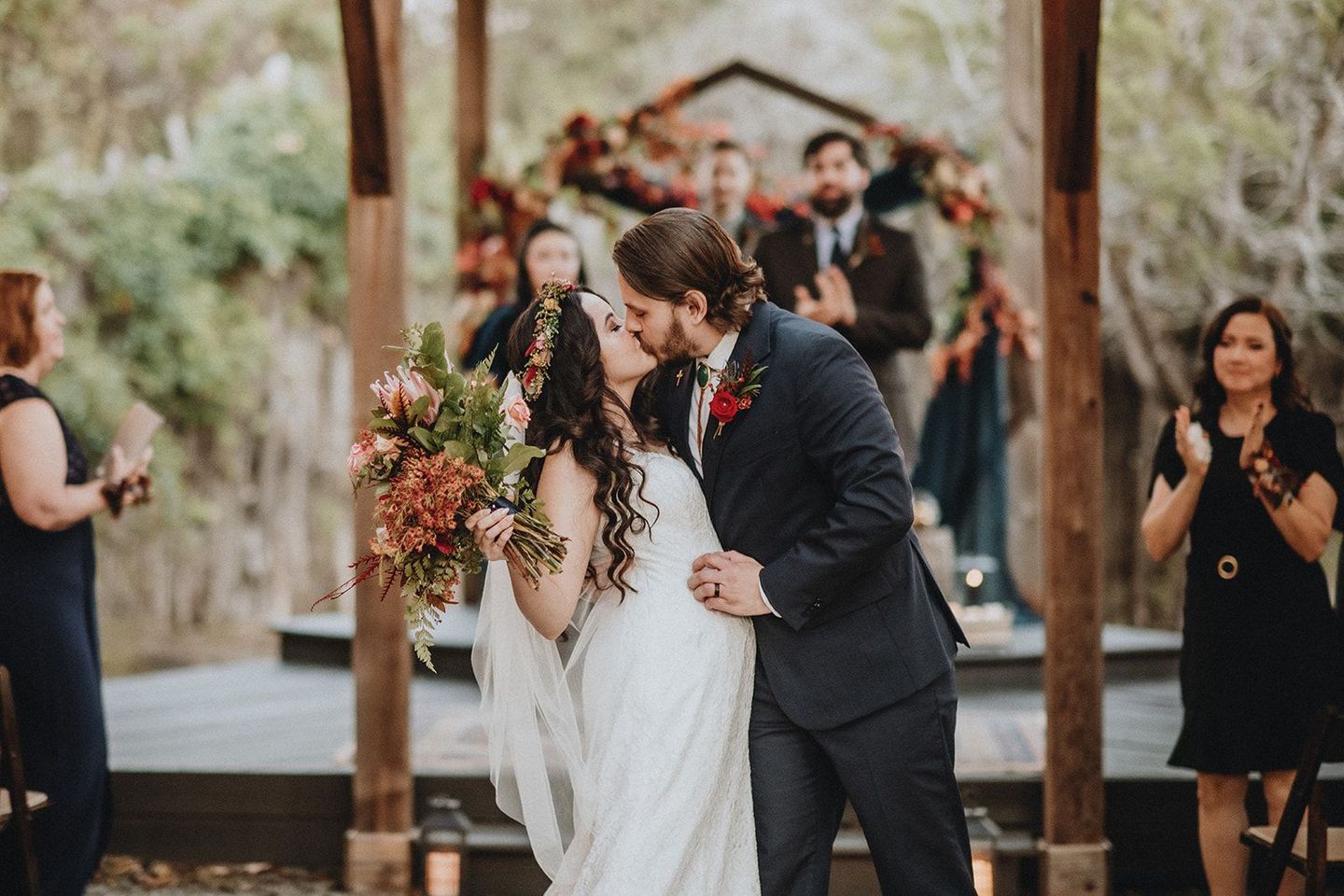 Wedding couple kissing beneath an arch, with guests applauding in a rustic outdoor ceremony