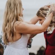 Two women embrace outdoors, one in a white dress and the other in red, in a tender moment.