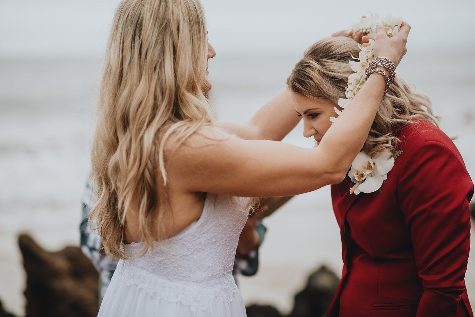 Woman placing a flower crown on another woman in a white dress outdoors by the water