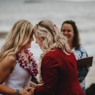 Two women embracing outdoors, one in a red jacket and the other in a white dress with a flower lei.
