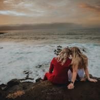 Two people sitting on a rocky shore, facing the ocean at sunset, one in a red dress.