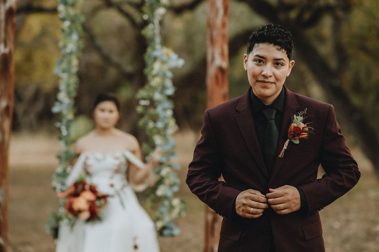 Bride and groom standing outdoors, with the groom in a burgundy suit and the bride holding a bouquet.